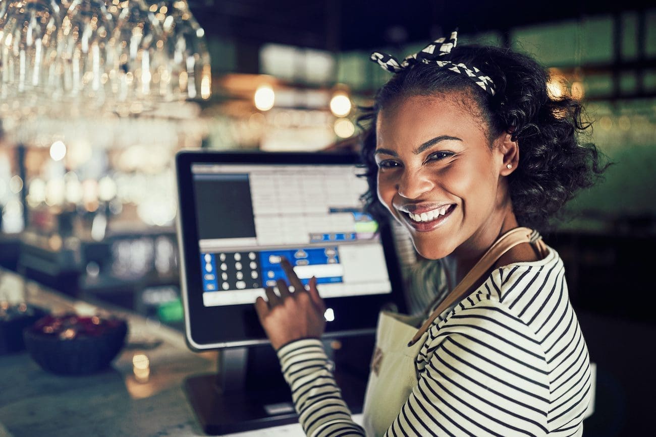 Smiling young African waitress wearing an apron using a touchscreen point of sale terminal while working in a trendy restaurant