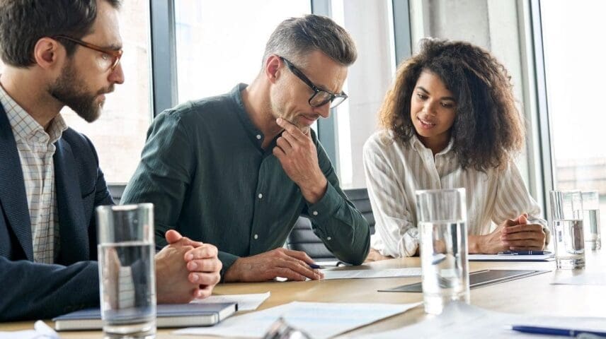 Focused doubtful mature businessman reading contract document thinking considering risks with professional lawyers legal experts executive team analyzing financial report sitting at office table.