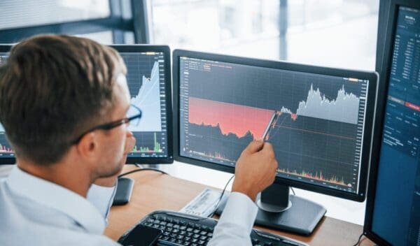 Looks at the graphs. Young businessman in formal clothes is in office with multiple screens. Conception of exchange and money.