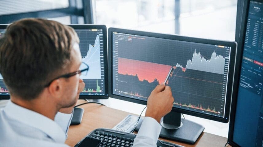 Looks at the graphs. Young businessman in formal clothes is in office with multiple screens. Conception of exchange and money.