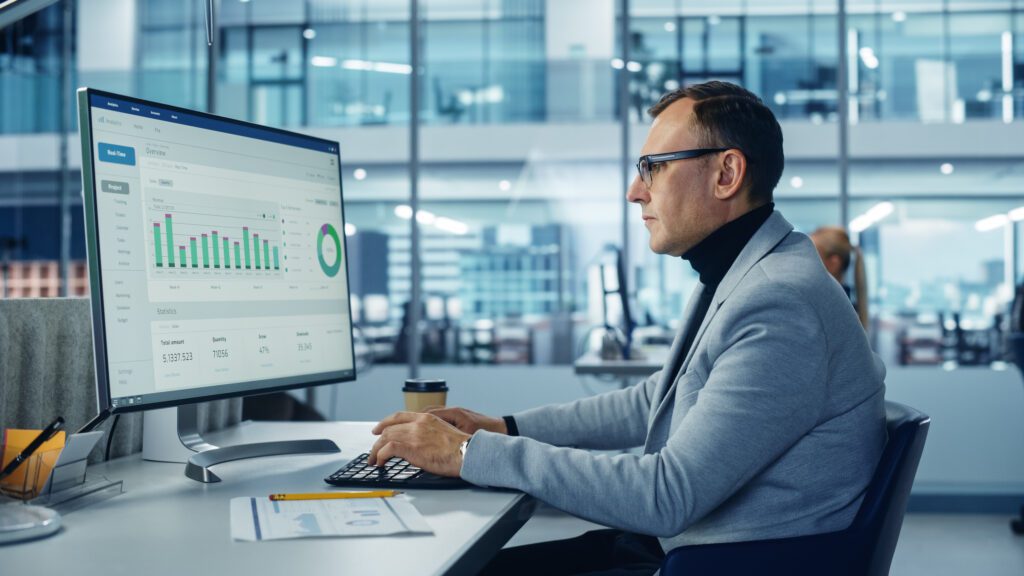 Diverse Corporate Office: Stylish Handsome Big Data Engineer Using Desktop Computer, Screen Showing Graphs, Statistics, Company Growth. Businessman Working on e-Commerce Project Marketing, Development