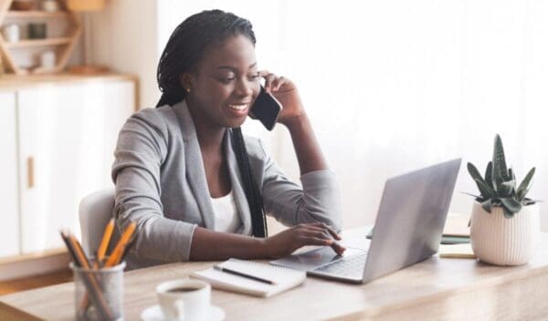 Busy Businesswoman Working At Desk In Office, Talking On Cellphone And Typing At Laptop, Copy Space