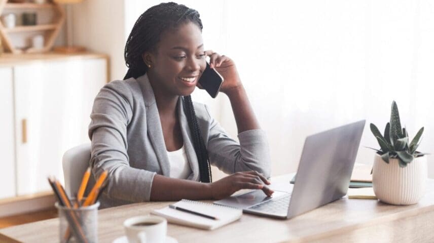 Busy Businesswoman Working At Desk In Office, Talking On Cellphone And Typing At Laptop, Copy Space