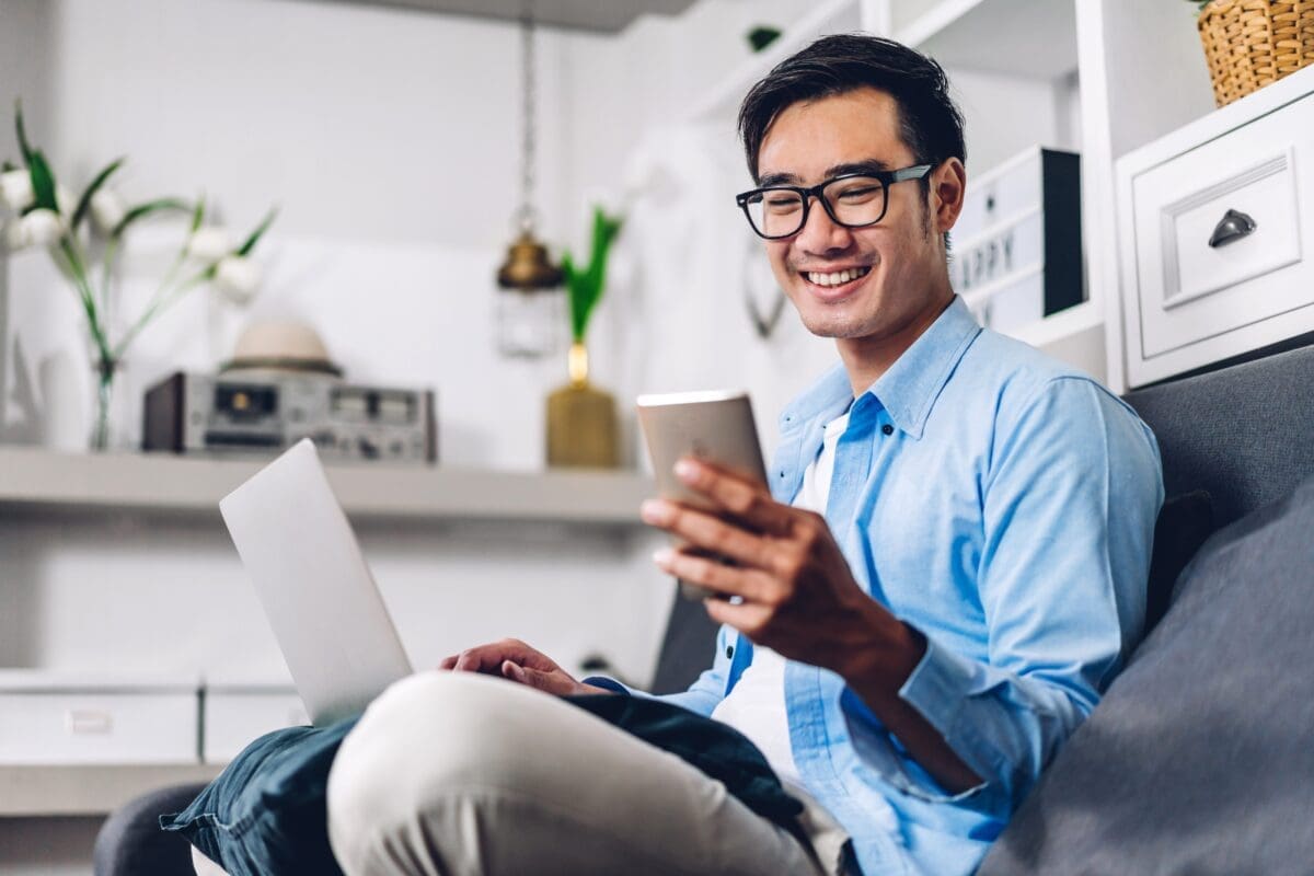 Young smiling asian man relaxing using laptop computer working and video conference meeting at home.Young creative man looking at screen typing message with smartphone.work from home concept