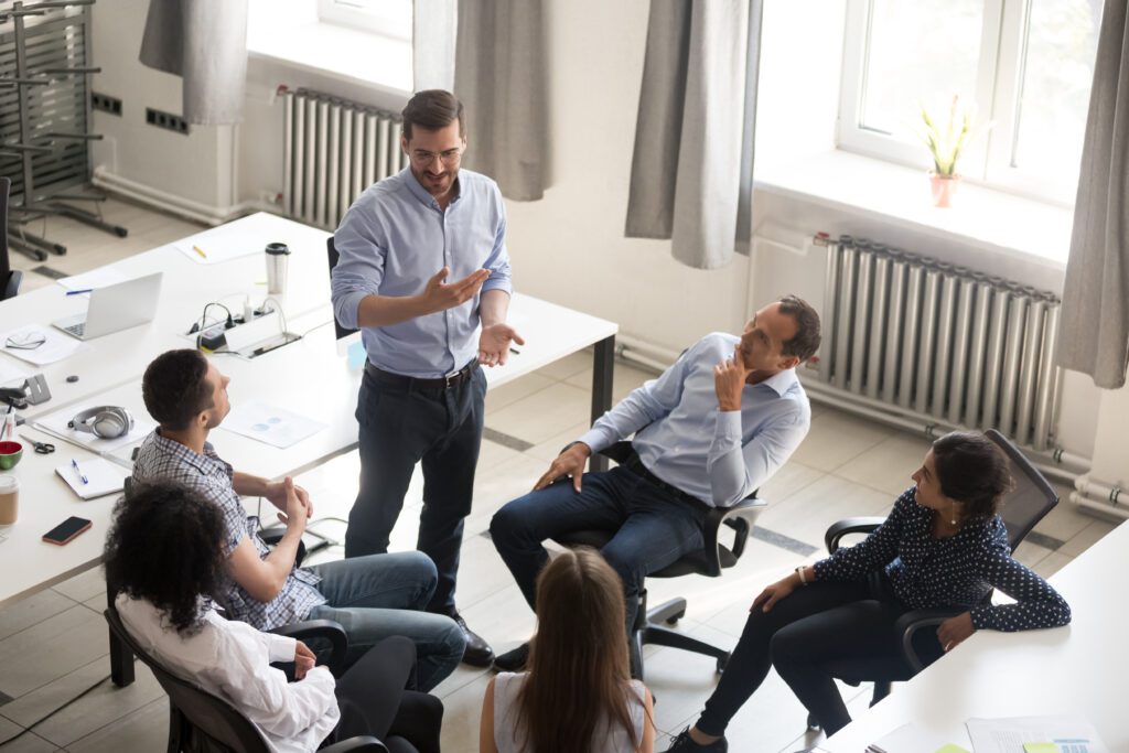 Smiling millennial male group leader stand in office talking with colleagues, discuss ideas, diverse employees brainstorm in shared space engaged in motivational teambuilding meeting with boss