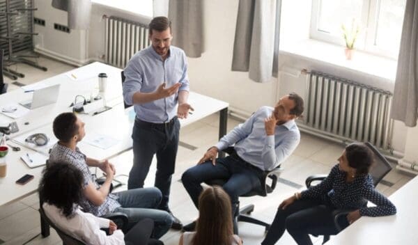 Smiling millennial male group leader stand in office talking with colleagues, discuss ideas, diverse employees brainstorm in shared space engaged in motivational teambuilding meeting with boss