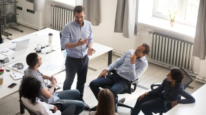 Smiling millennial male group leader stand in office talking with colleagues, discuss ideas, diverse employees brainstorm in shared space engaged in motivational teambuilding meeting with boss