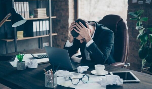 Photo of helpless business guy crumple papers holding head hands, need rest work day night workaholic tired powerless wear black blazer shirt suit sitting chair office indoors