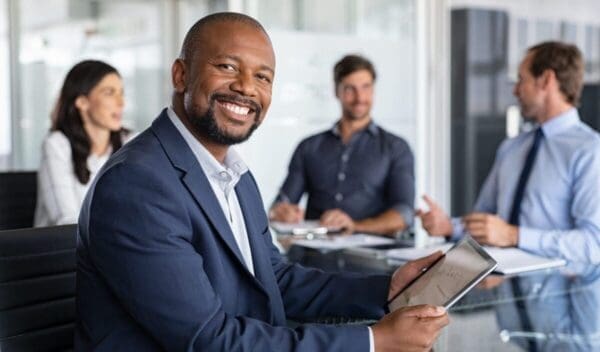 Mature black businessman with colleagues sitting in a modern board room. Proud smiling business man sitting during a meeting and looking at camera. Portrait of happy successful executive with team working in background.