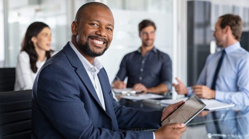 Mature black businessman with colleagues sitting in a modern board room. Proud smiling business man sitting during a meeting and looking at camera. Portrait of happy successful executive with team working in background.