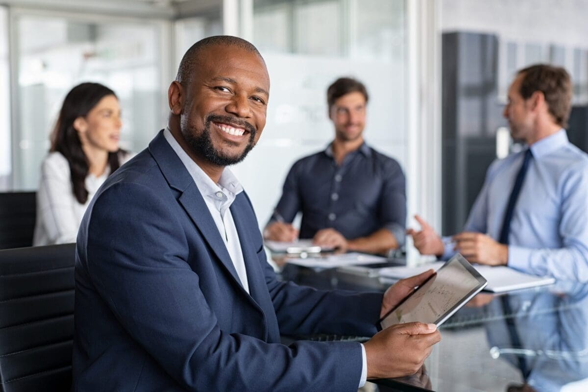 Mature black businessman with colleagues sitting in a modern board room. Proud smiling business man sitting during a meeting and looking at camera. Portrait of happy successful executive with team working in background.